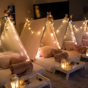 Indoor sleepover setup with small teepee tents, floor mattresses, pillows, blankets, fairy lights, and tray tables.